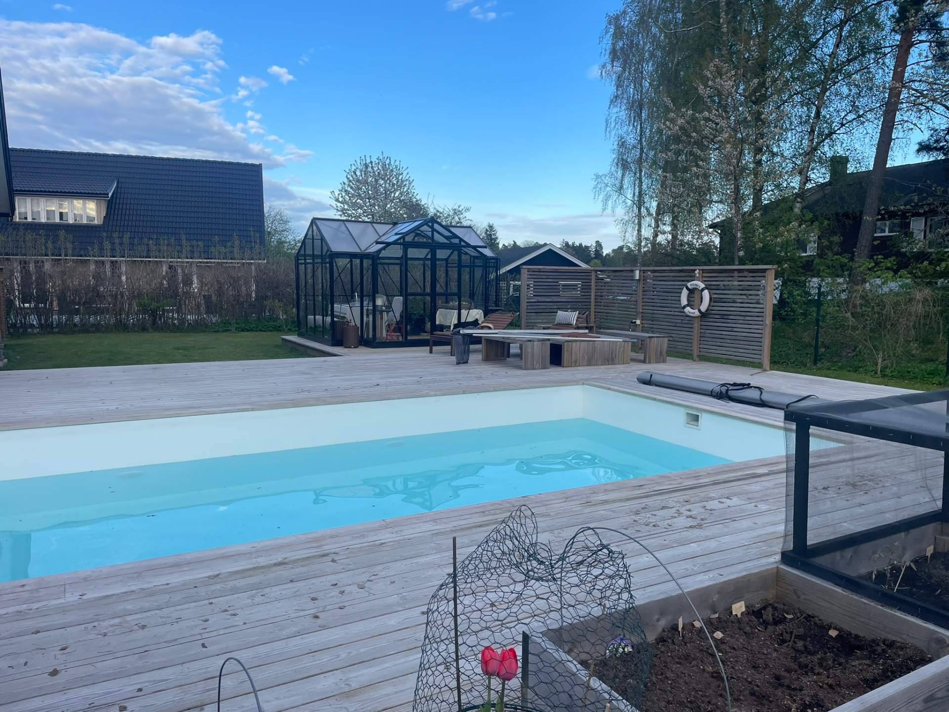 Färdig pool i trädgården — trall, växthus, insynsskydd i ribbat trä och livboj